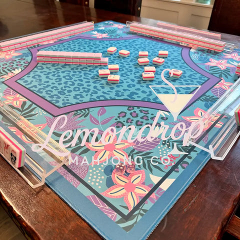 A colorful mahjong mat with a blue leopard print center, pink and purple borders, and tropical floral patterns, shown on a dark wooden table with several pink-backed tiles arranged on top.