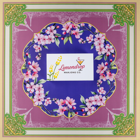 Square decorative mat design featuring a deep purple circular center framed by pink cherry blossom branches with green leaves. In the center is the Lemondrop Mahjong Co. tile box with logo, yellow flowers and a martini glass icon. The outer border is dusty pink with fine white line illustrations of Paris landmarks, surrounded by a gold and green ornamental frame with floral corner details, giving an elegant Parisian, floral aesthetic.